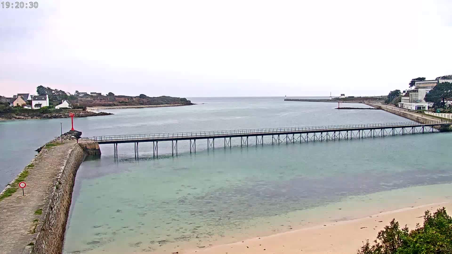 A long pier stretches across calm, shallow, light-green water towards a small island under an overcast sky.
