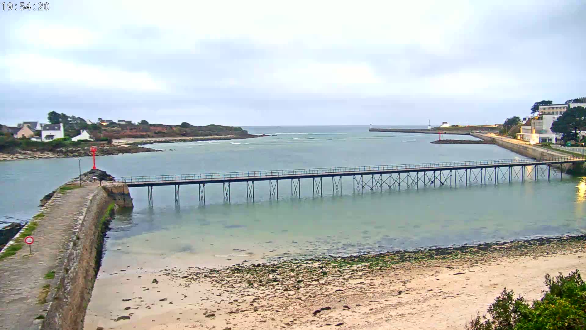 A long pier stretches across calm, shallow water separating a sandy beach from a small town under an overcast sky.