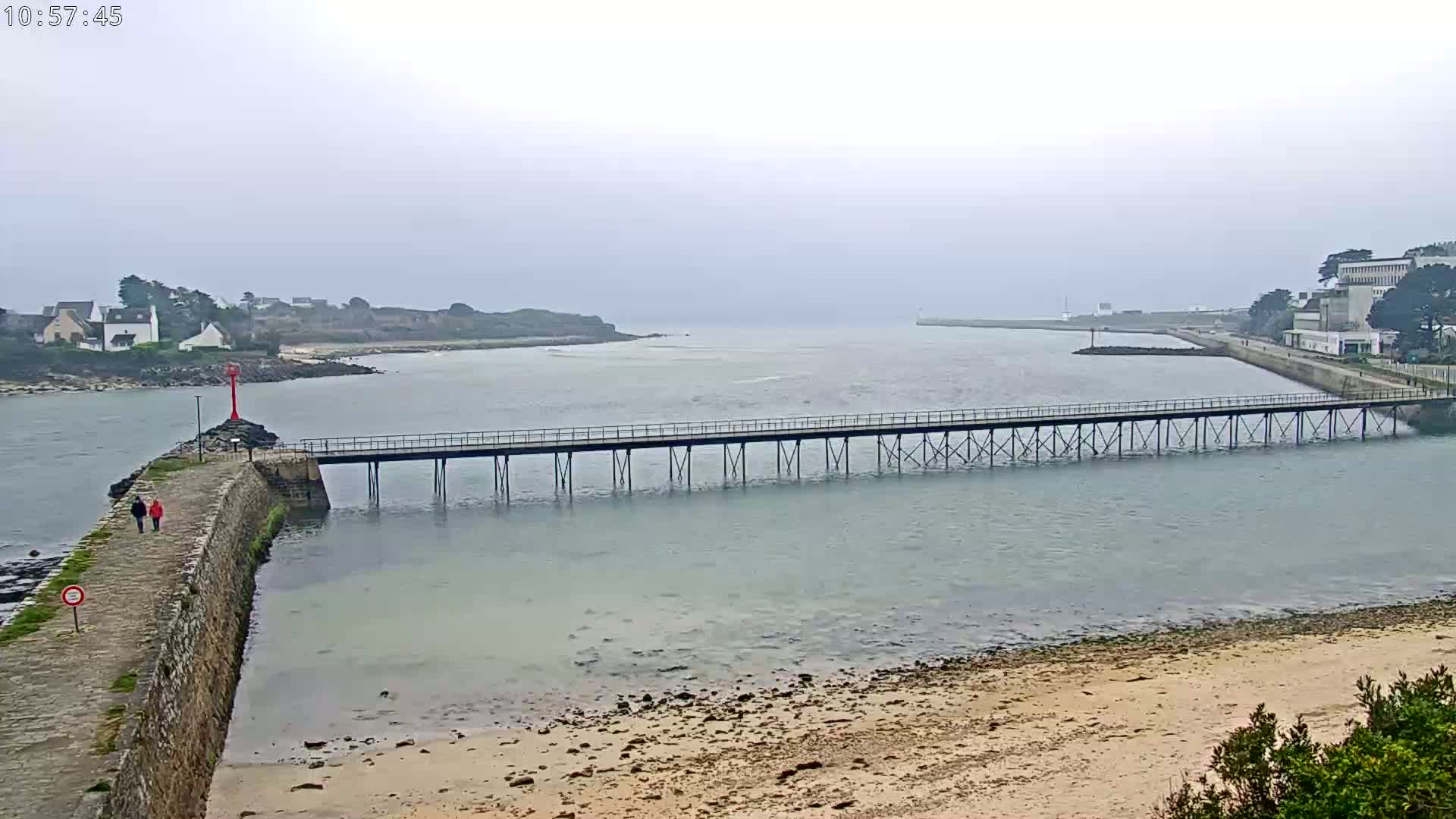 A long pier stretches across calm, light-green water under an overcast sky, connecting a stone wall to a distant shore with buildings.