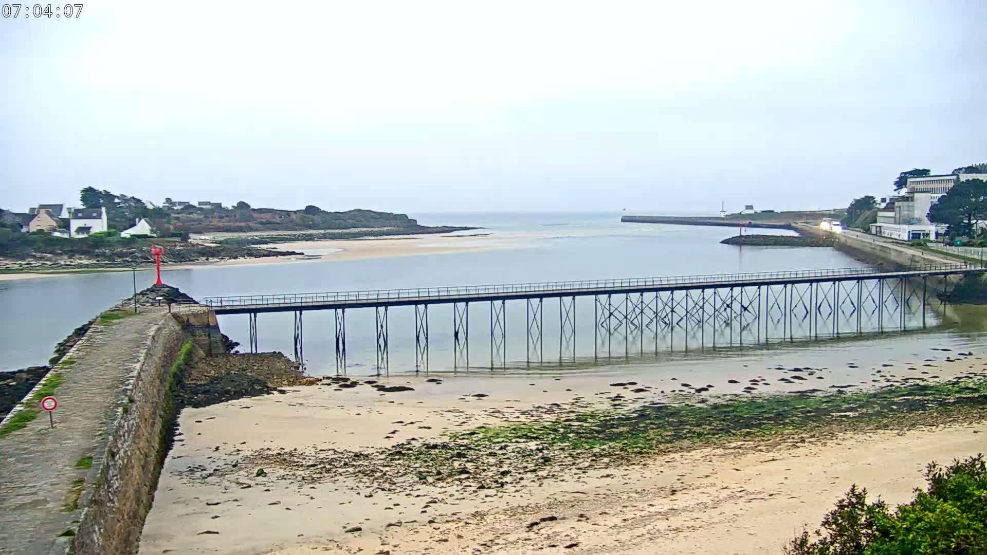 A long pier stretches across calm, shallow water toward a small town on a cloudy day, with sandy beaches and rocky shorelines visible on either side.