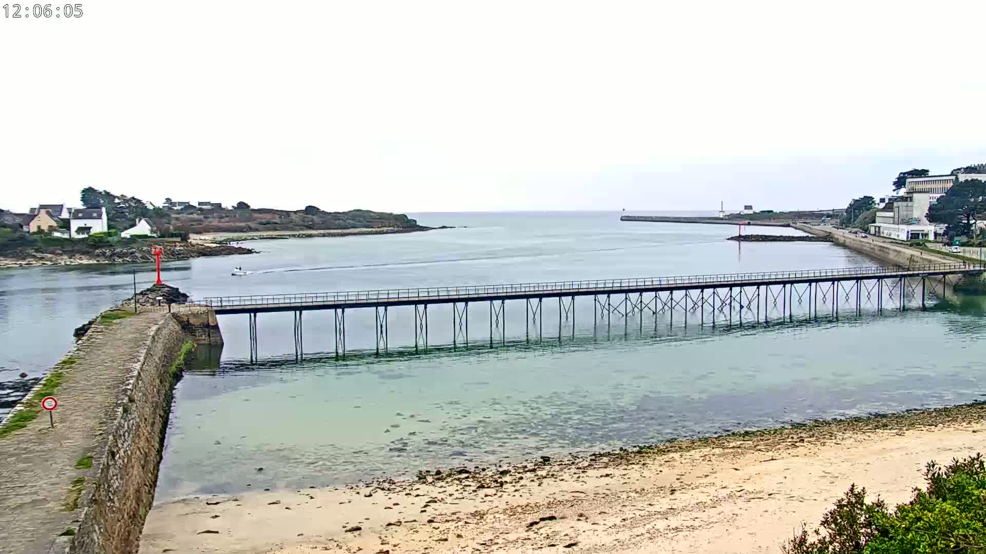 A long pier extends over calm, shallow water toward a small island and a distant shoreline under an overcast sky.