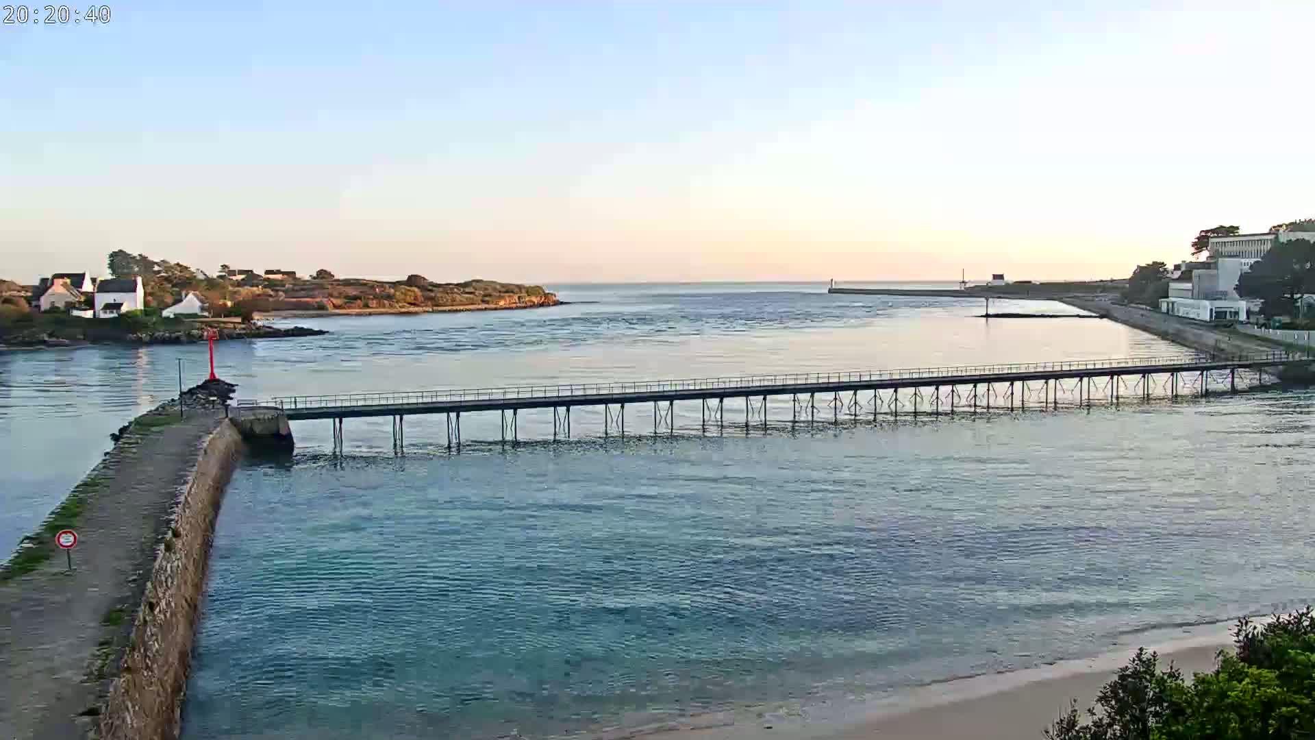 A long pier stretches across calm, blue-green water toward a small island under a clear, pale sunrise sky.