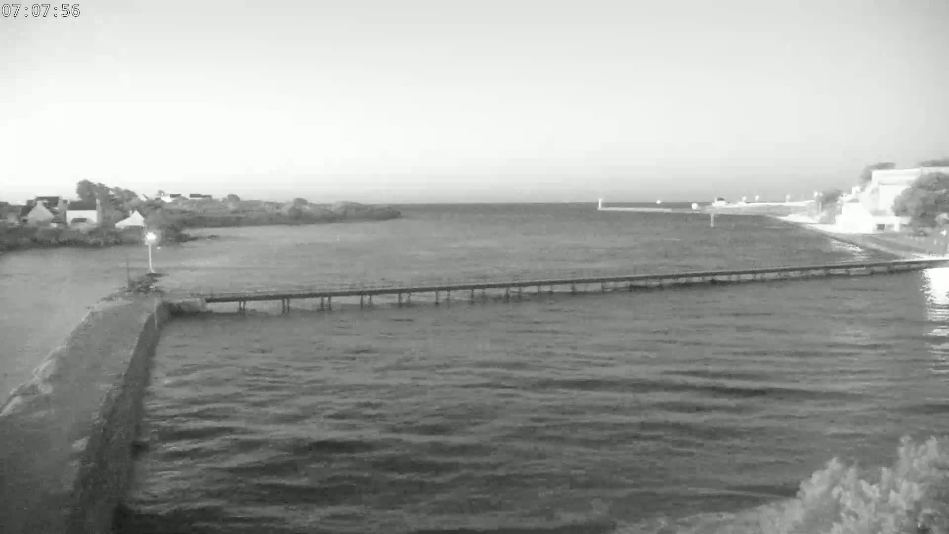 A long pier extends over a calm body of water, with a small town visible in the background under a light overcast sky.