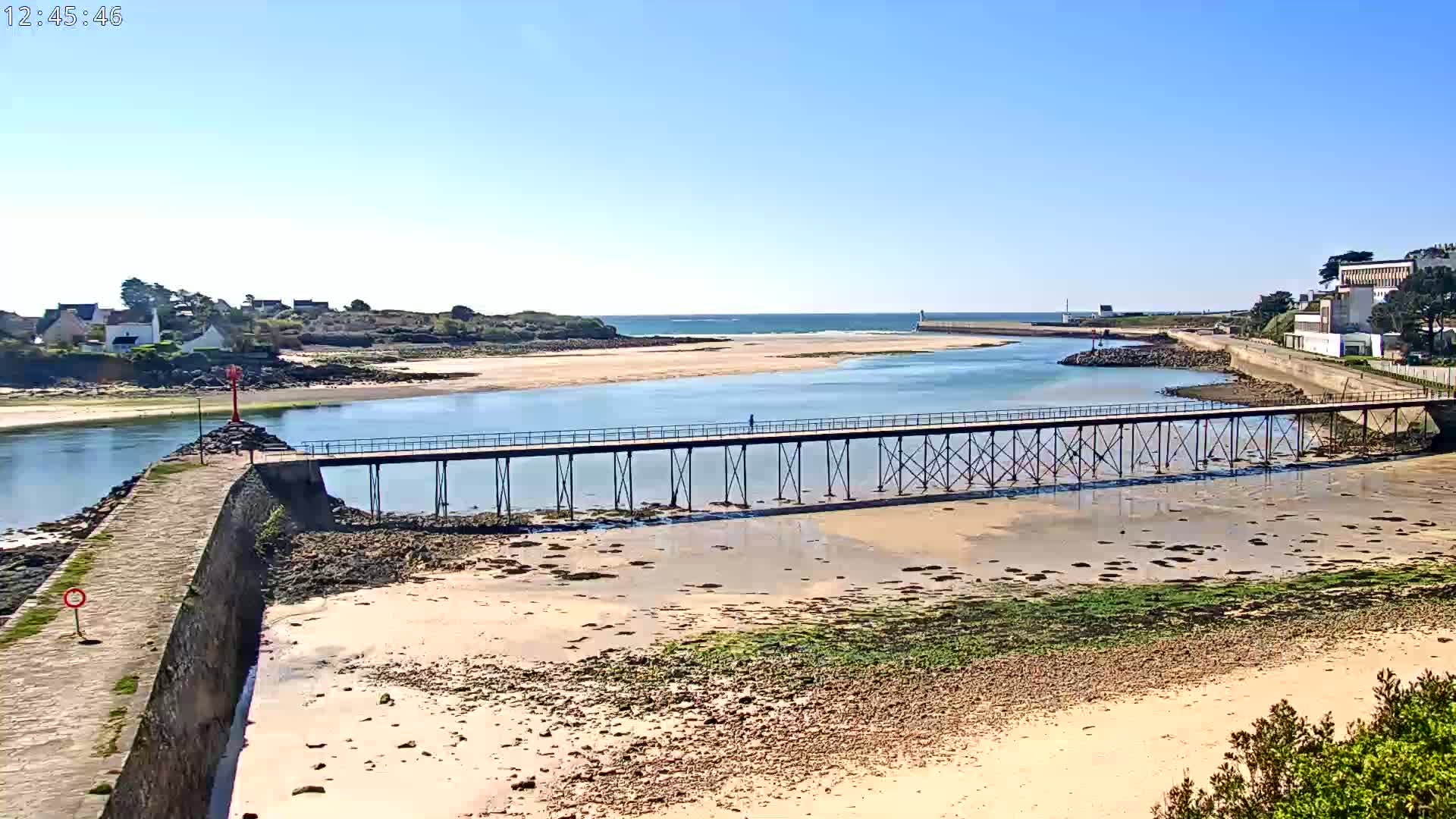 A long pier stretches across a calm body of water, connecting a sandy beach to a small coastal town on a sunny day.