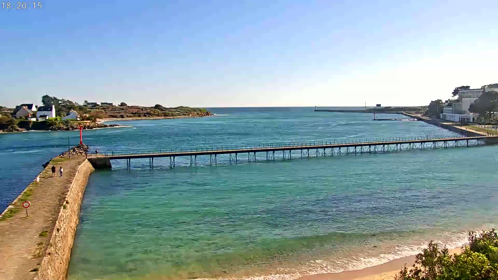 A long pier extends across calm, clear turquoise water toward a small island under a bright, sunny sky.