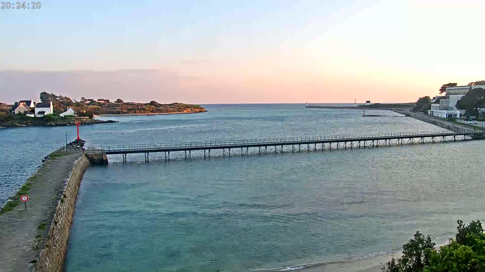 A long pier extends across calm, light-blue water at sunset, connecting a stone jetty to a distant landmass with buildings.
