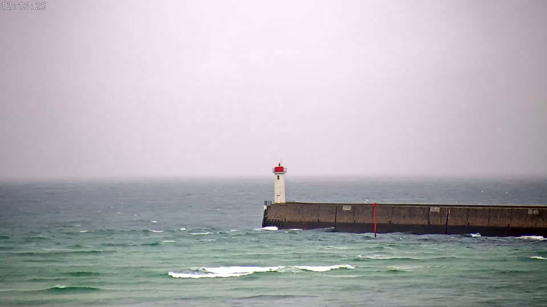 A white lighthouse with a red top sits atop a long, dark breakwater in a gray, overcast ocean with small waves.
