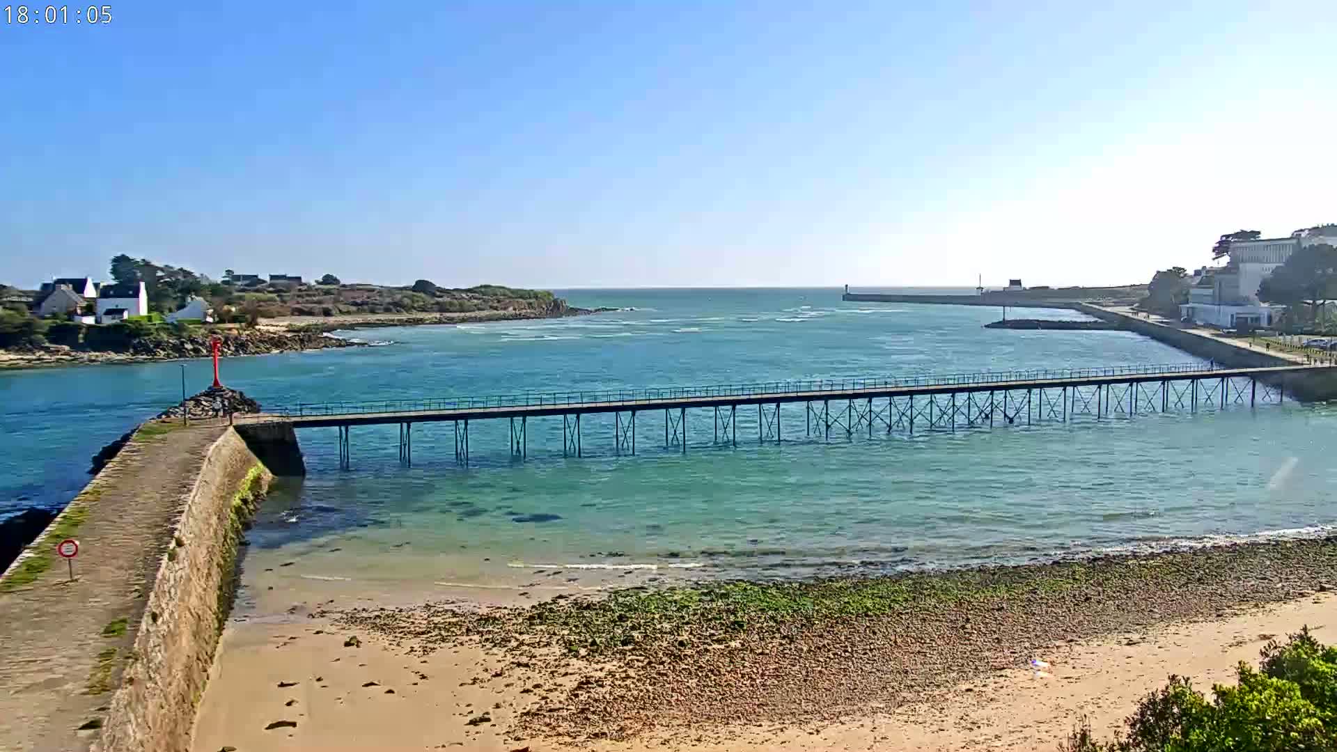 A long pier extends over calm, teal water toward a small, distant island under a clear blue sky.