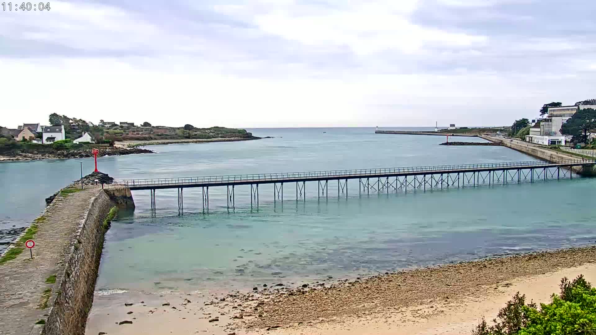 A long pier stretches across calm, light-green water, connecting a stone jetty to a landmass with buildings, under an overcast sky.