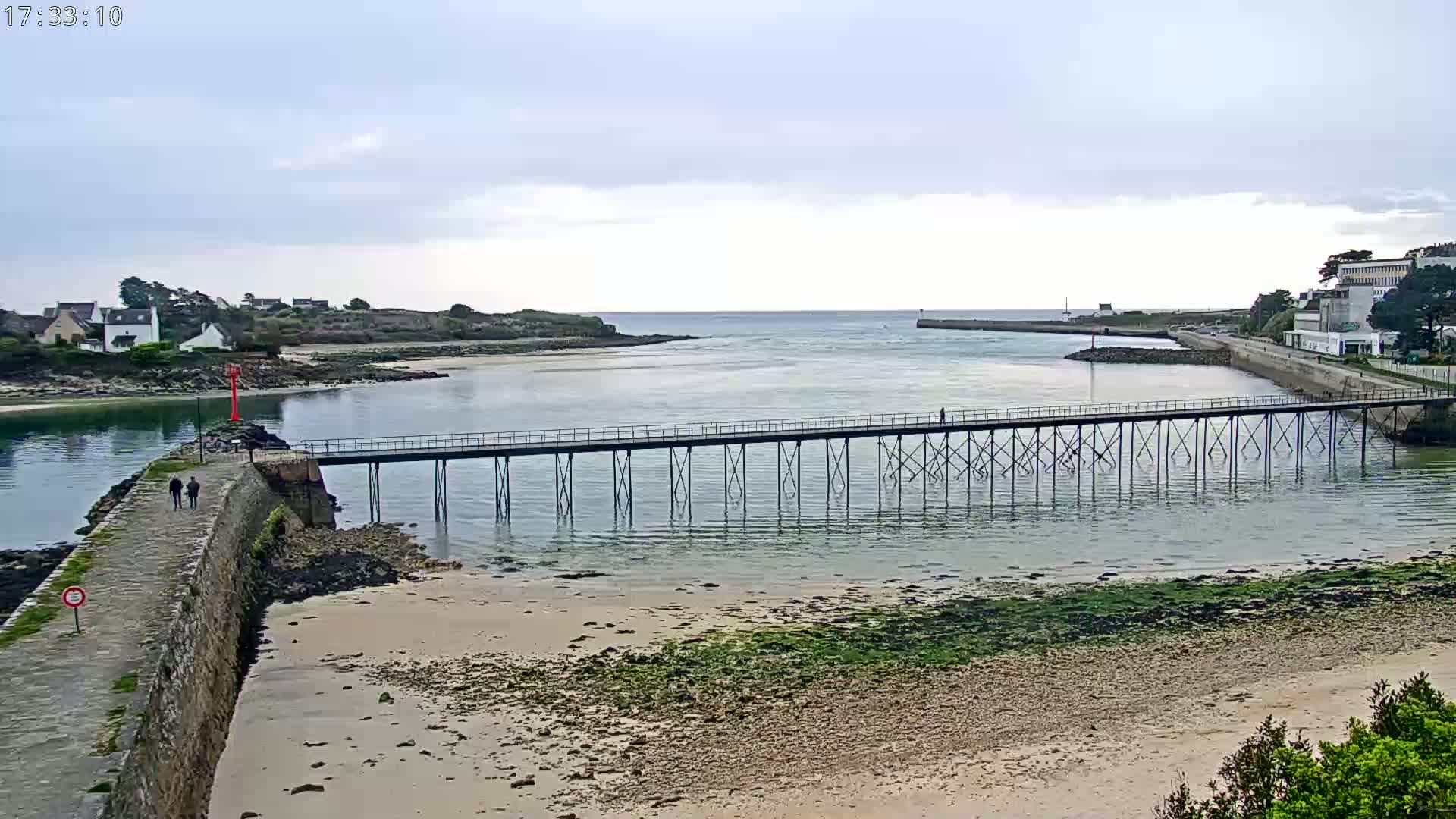 A long pier stretches across calm, shallow water between two shorelines on an overcast day, with a sandy beach visible below.