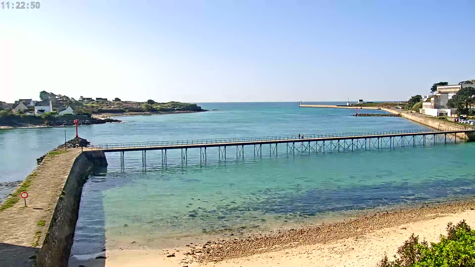 A long pier stretches across clear, turquoise water toward a small landmass and a town under a bright, sunny sky.