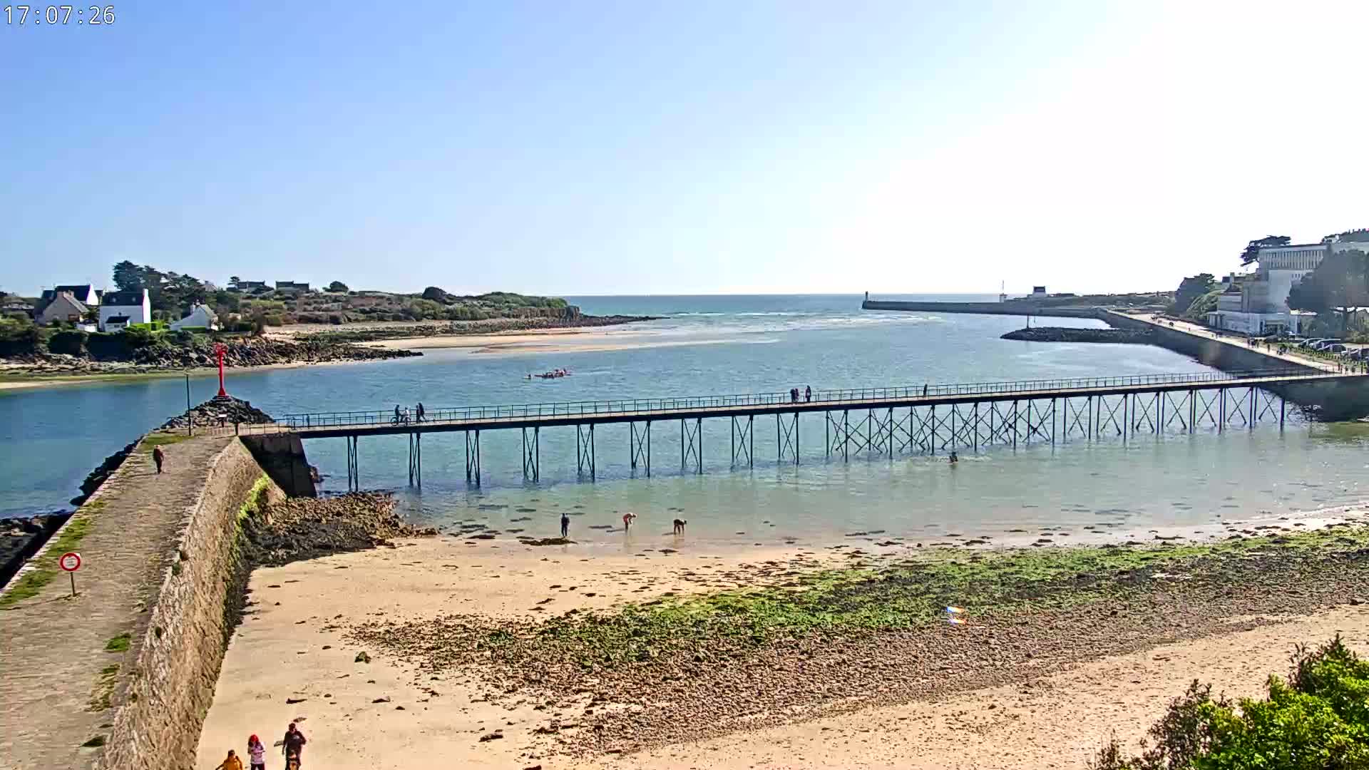 A long pier extends over calm, shallow water toward a small island on a sunny day, with people walking on the pier and some wading in the water near a sandy beach.