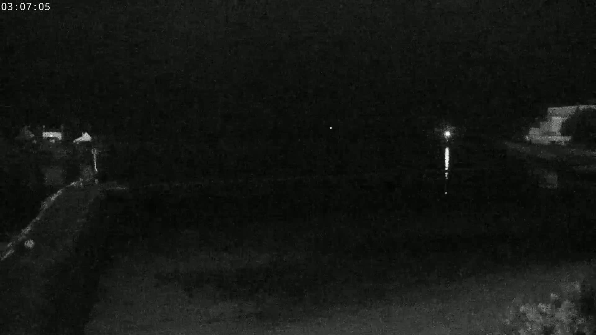 A nighttime, low-light view shows a dark body of water reflecting a distant light source, with dimly lit structures visible on the shore.