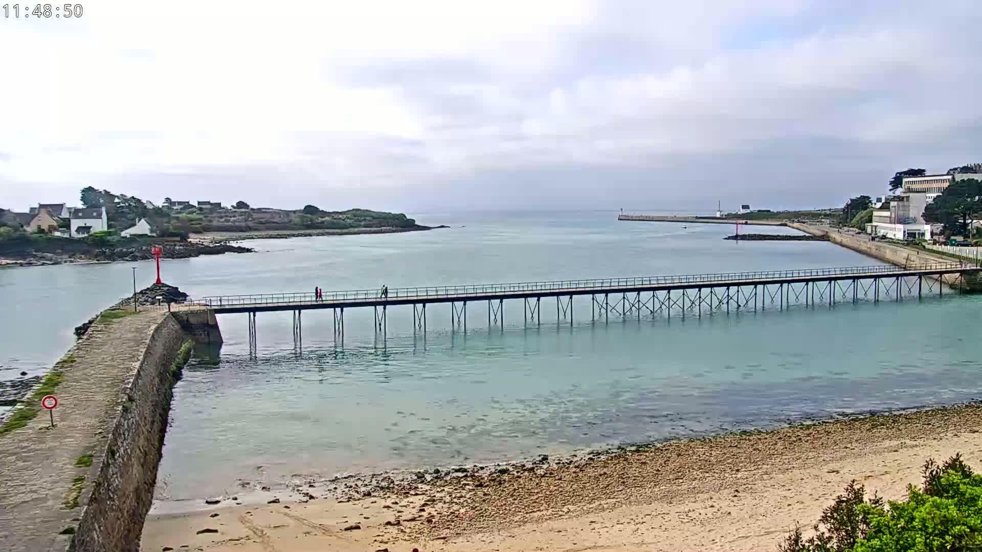 A long pier stretches across calm, light-blue water toward a small town under a mostly cloudy sky.