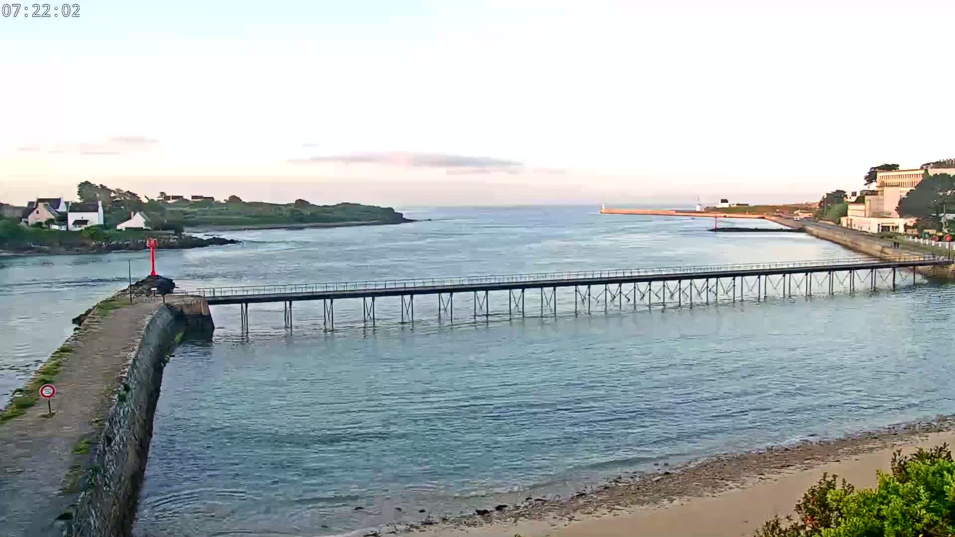 A long pier extends over calm, light-blue water toward a small town and island under a pale, mostly clear sky.