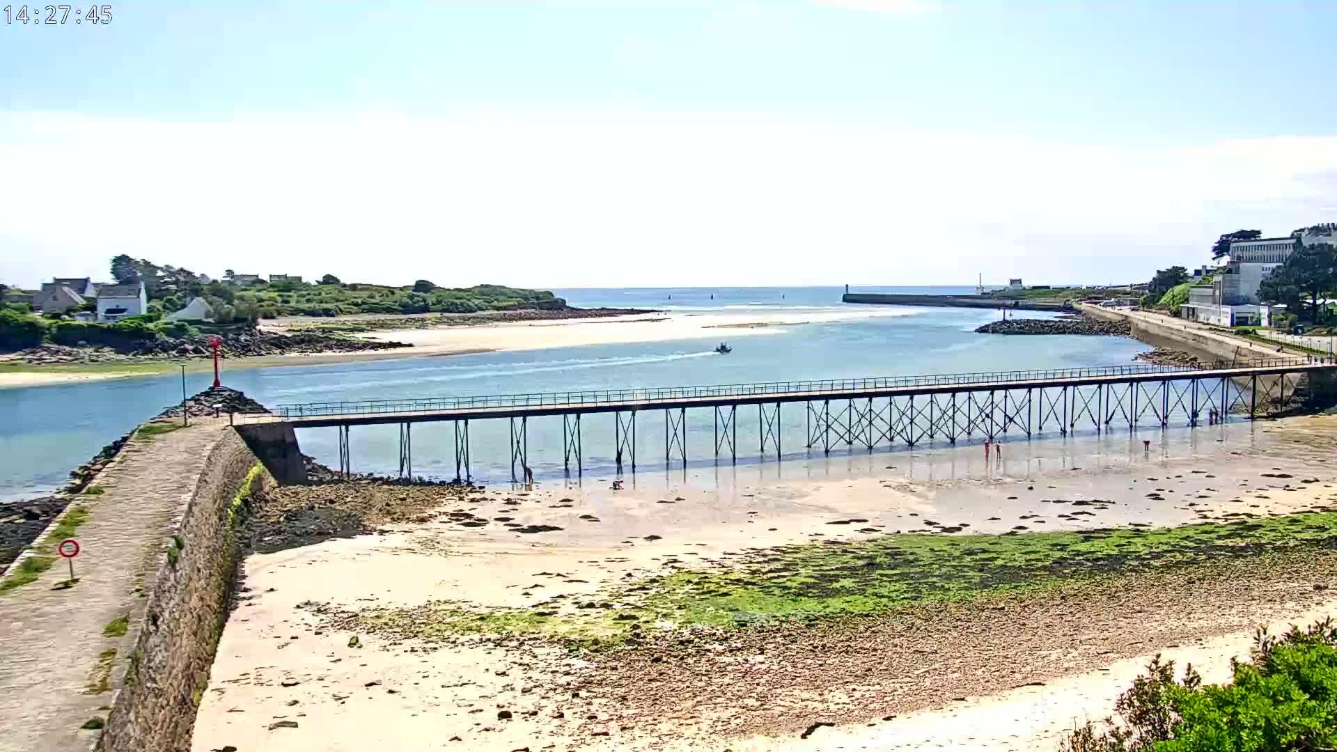 A long pier extends over a calm, shallow waterway leading to a sandy beach, under a bright, sunny sky.