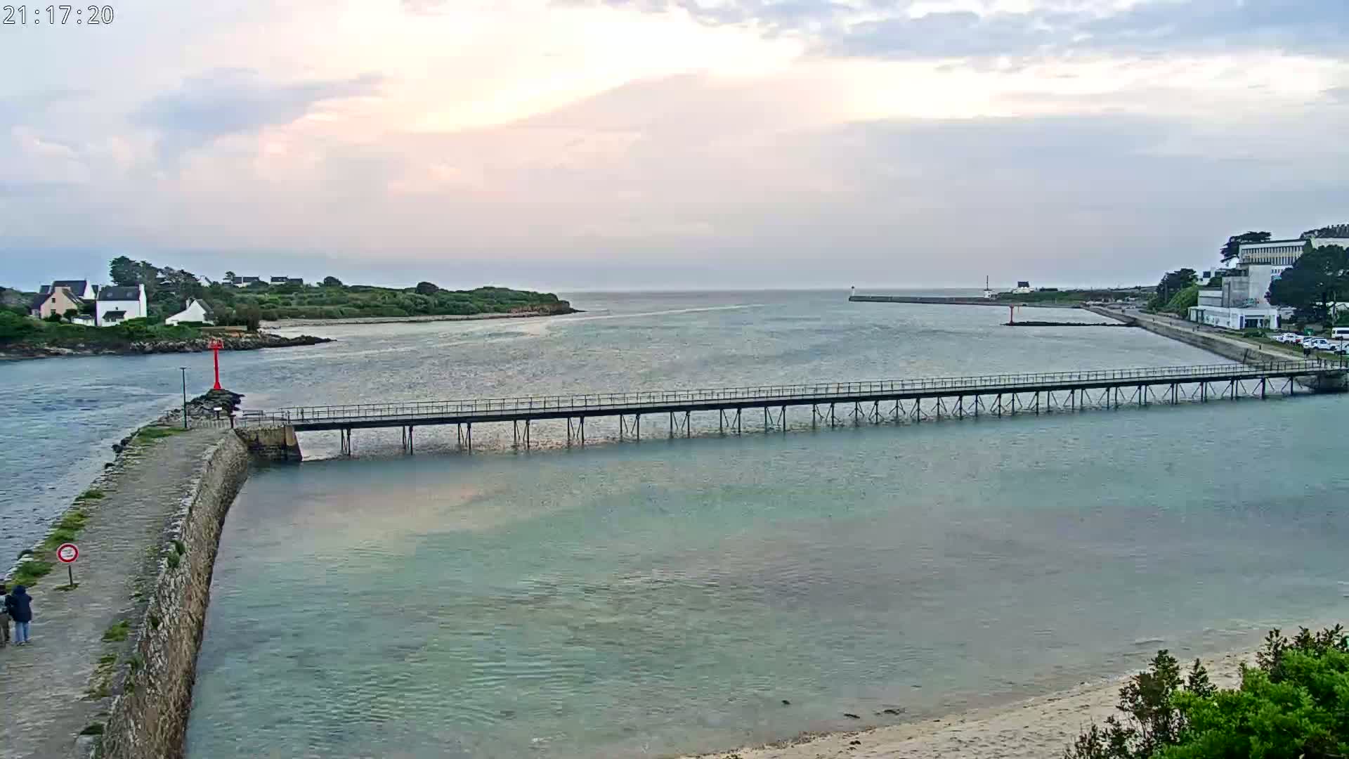 A long pier stretches across a calm body of water under a cloudy sky, connecting a stone jetty to a landmass with buildings in the distance.