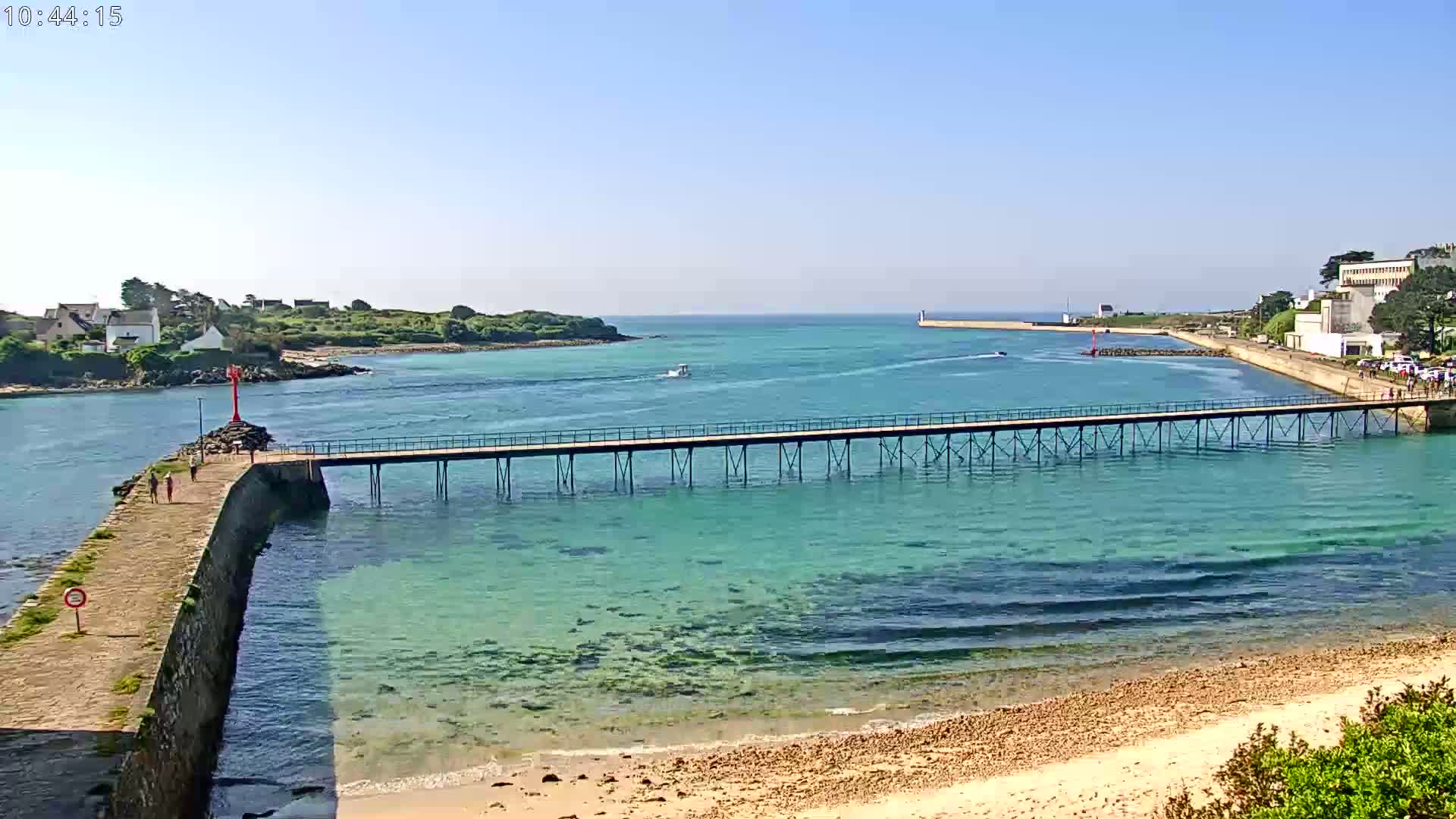 A long pier stretches across clear, turquoise water toward a distant shoreline on a sunny day, with small boats visible in the distance.