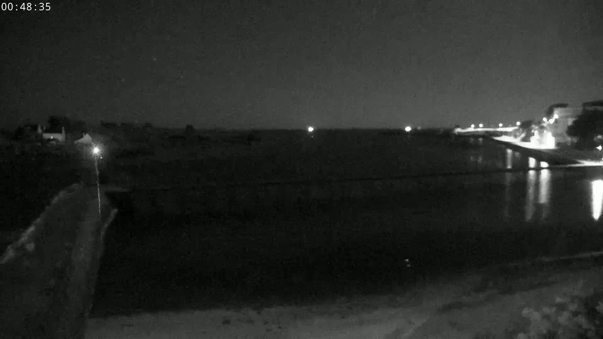A nighttime view shows a dark body of water reflecting lights from a distant shoreline under a clear, starlit sky.