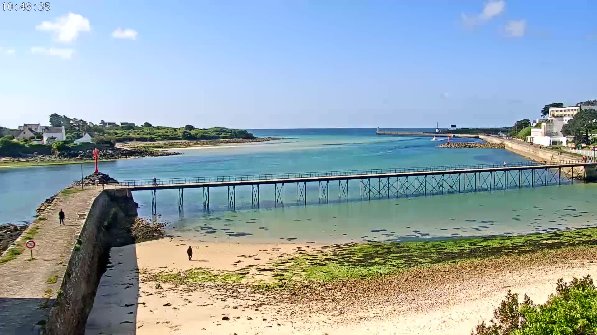 A long pier stretches across shallow, clear turquoise water toward a small island under a bright, sunny sky.