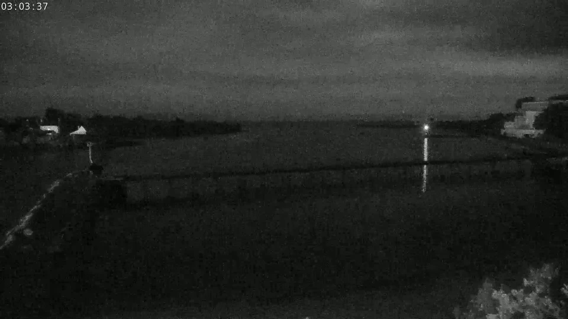 A nighttime, low-light view shows a dark body of water reflecting a single light source on the far shore, with dark land and buildings visible in the foreground and background.