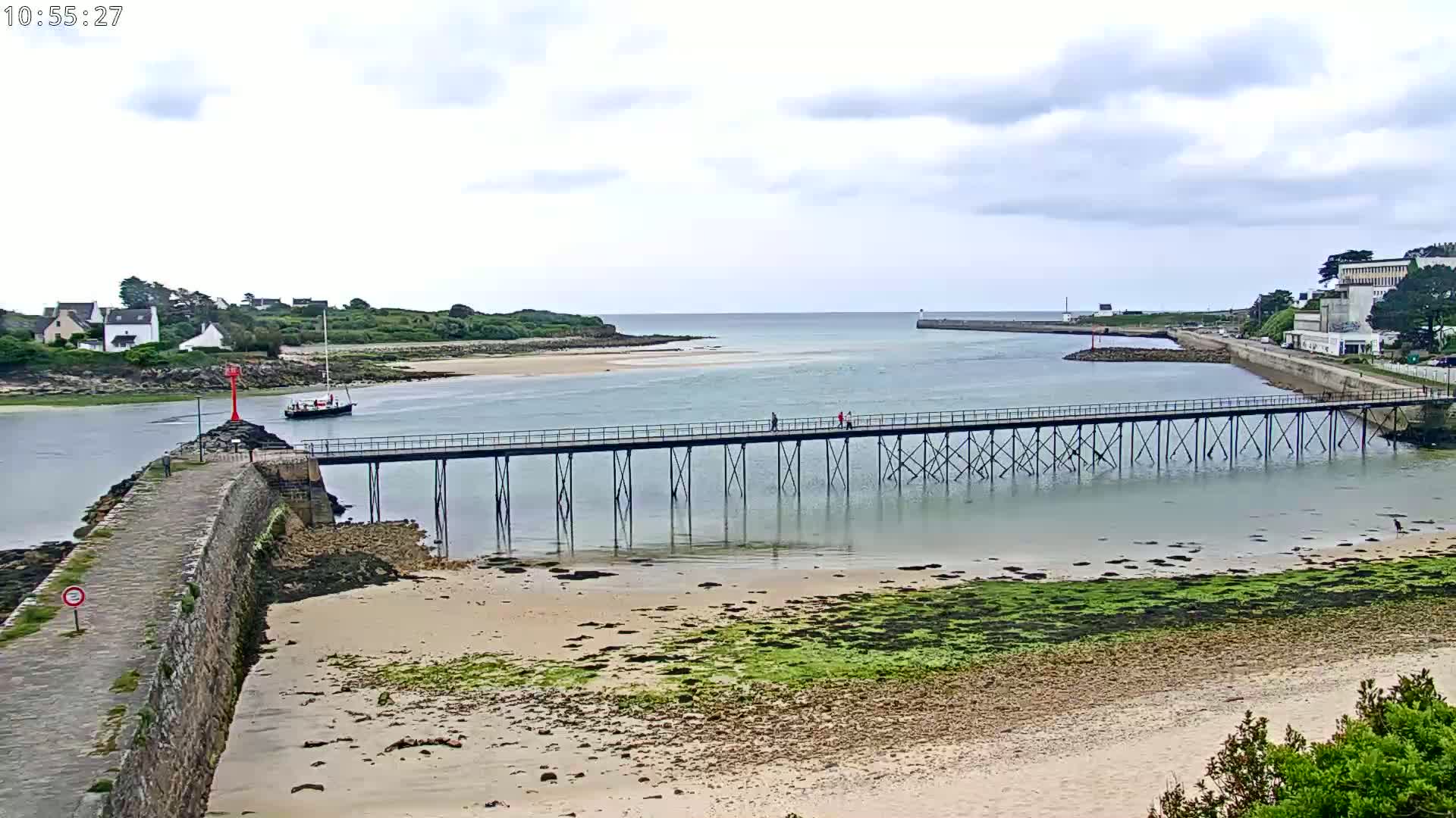 A long pier stretches across calm, shallow water toward a small boat and a shoreline with buildings under a cloudy sky.