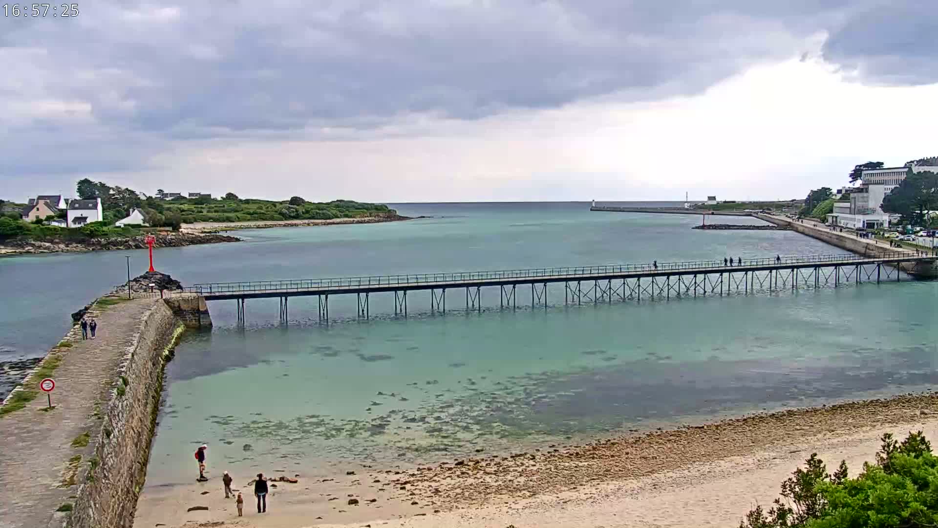 A long pier stretches across calm, shallow, teal water separating a sandy beach from a stone walkway leading to a small village under a cloudy sky.