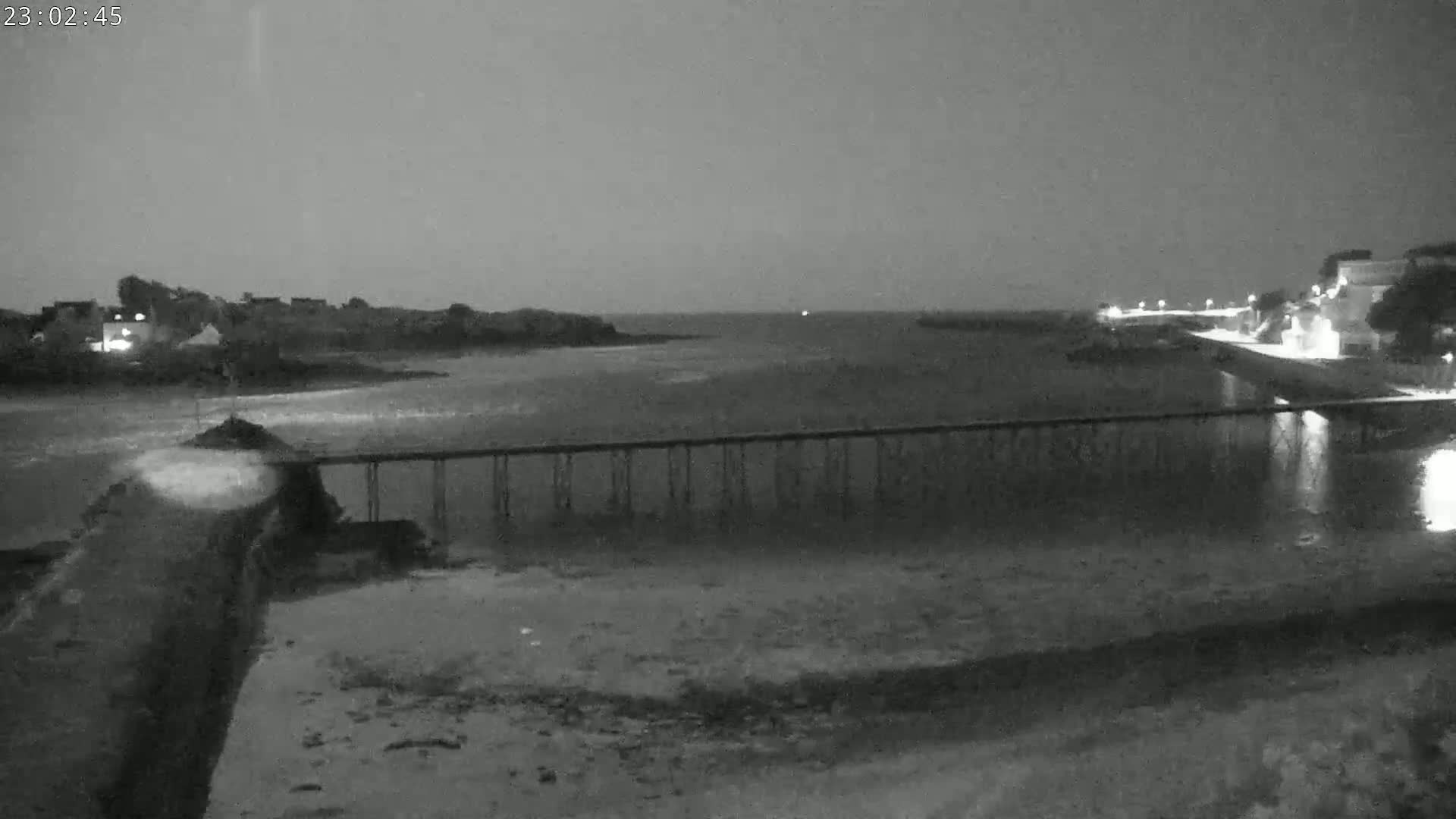 A long pier stretches across a dark, calm body of water at night, with faintly lit buildings visible on the far shore.