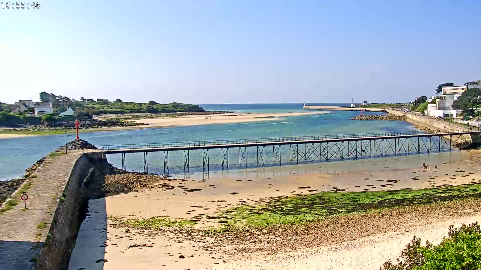 A long pier extends over shallow, sandy water, connecting a rocky shoreline to a breakwater under a clear, sunny sky.
