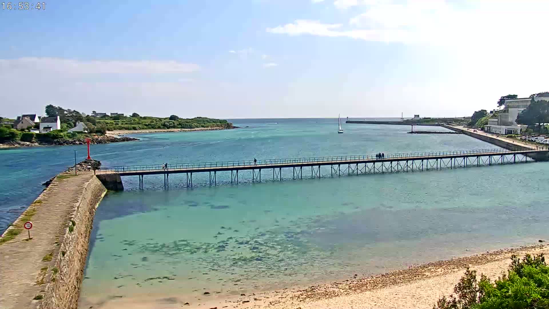 A long pier extends over clear, shallow turquoise water toward a small island under a partly sunny sky.
