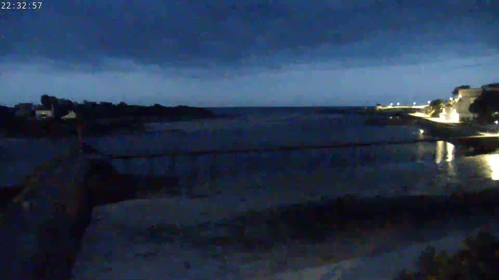A dark, calm body of water is seen at night, with a dimly lit shoreline and buildings visible in the distance under a mostly cloudy sky.