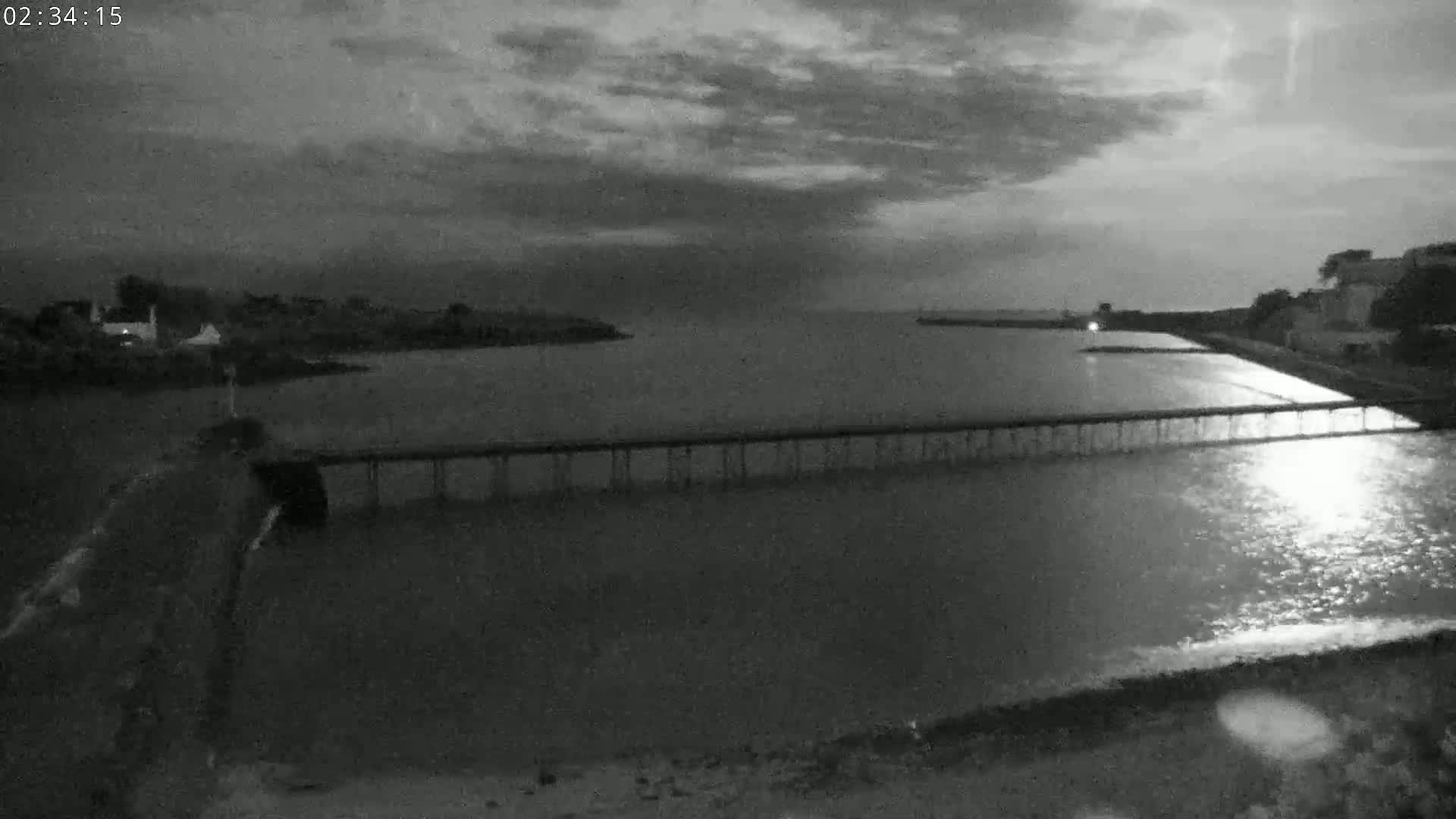 A long pier stretches across a calm body of water under a cloudy night sky.