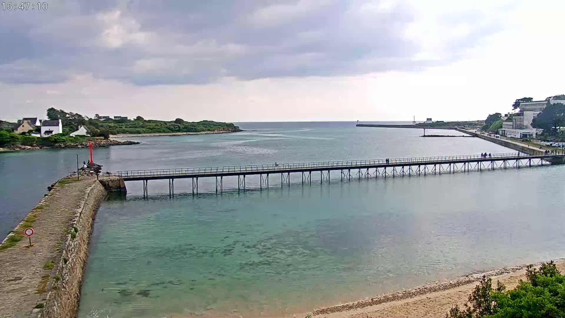 A long pier stretches across calm, clear water between two shorelines under a cloudy sky.