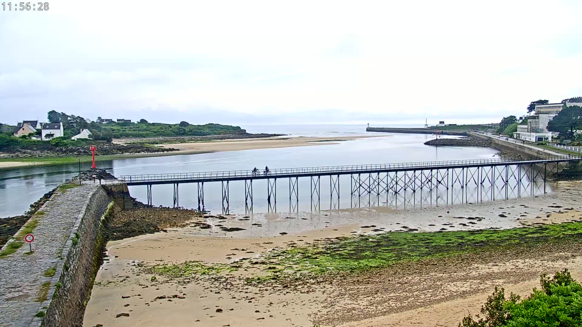 Two people ride bicycles across a long pier extending over calm water and a sandy shore under an overcast sky.