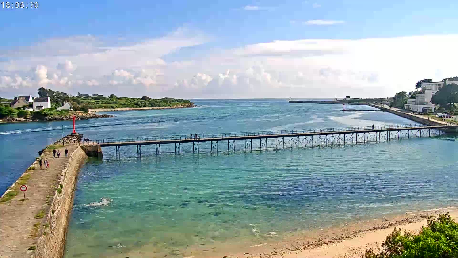 A long pier extends across calm, clear turquoise water, under a partly cloudy blue sky, toward a small town on a peninsula.
