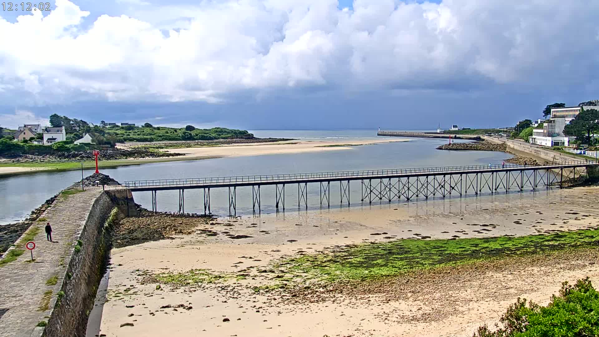 A long pier stretches across a calm body of water under a partly cloudy sky, with a sandy shore and low-lying green vegetation visible in the foreground.