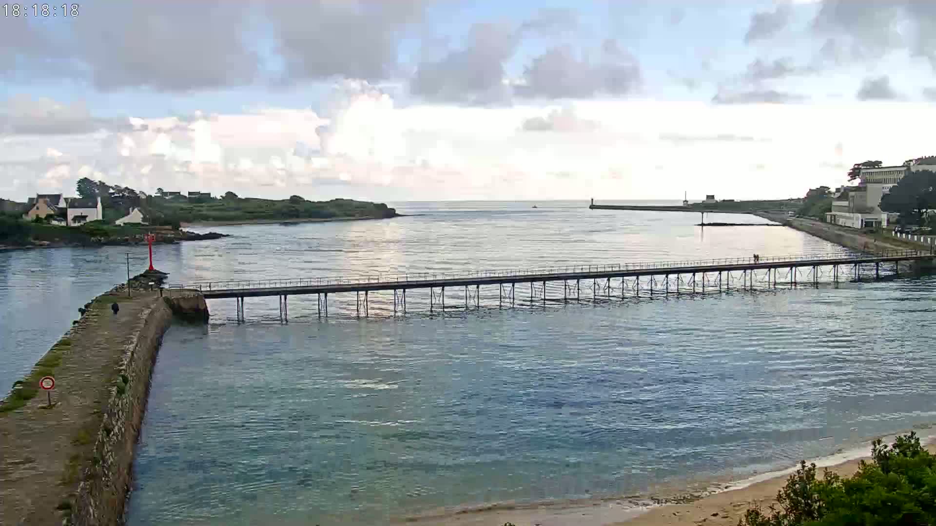 A long pier stretches across calm, blue water under a partly cloudy sky, connecting a stone jetty to a shoreline with buildings and green vegetation.