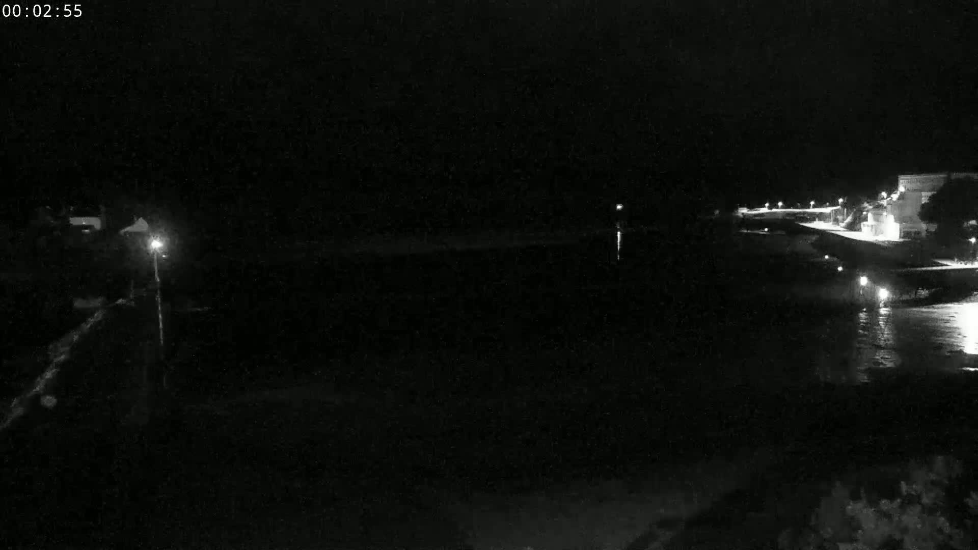 A dark, nighttime scene shows a body of water reflecting dim lights from a shoreline with buildings.