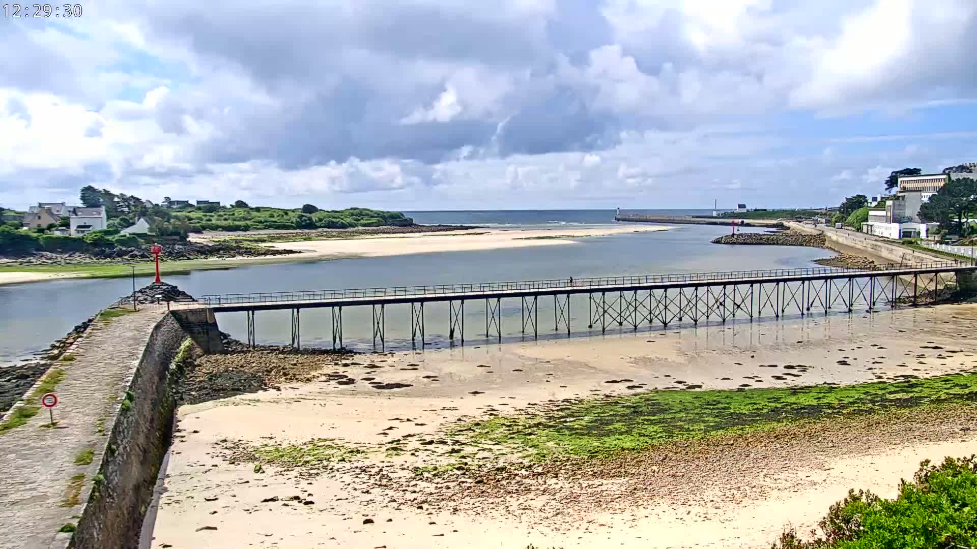 A long pier stretches across a calm bay with a sandy bottom partially exposed at low tide under a partly cloudy sky.