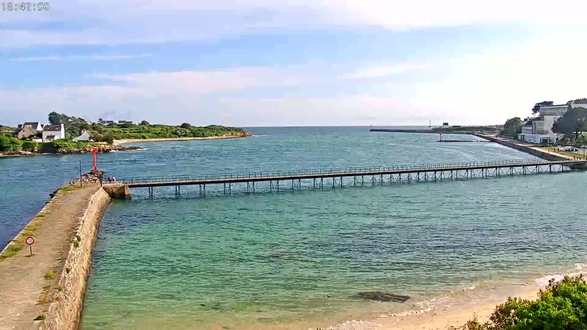 A long pier stretches across calm, teal water, connecting a stone jetty to a shoreline with buildings and green land under a mostly sunny sky.