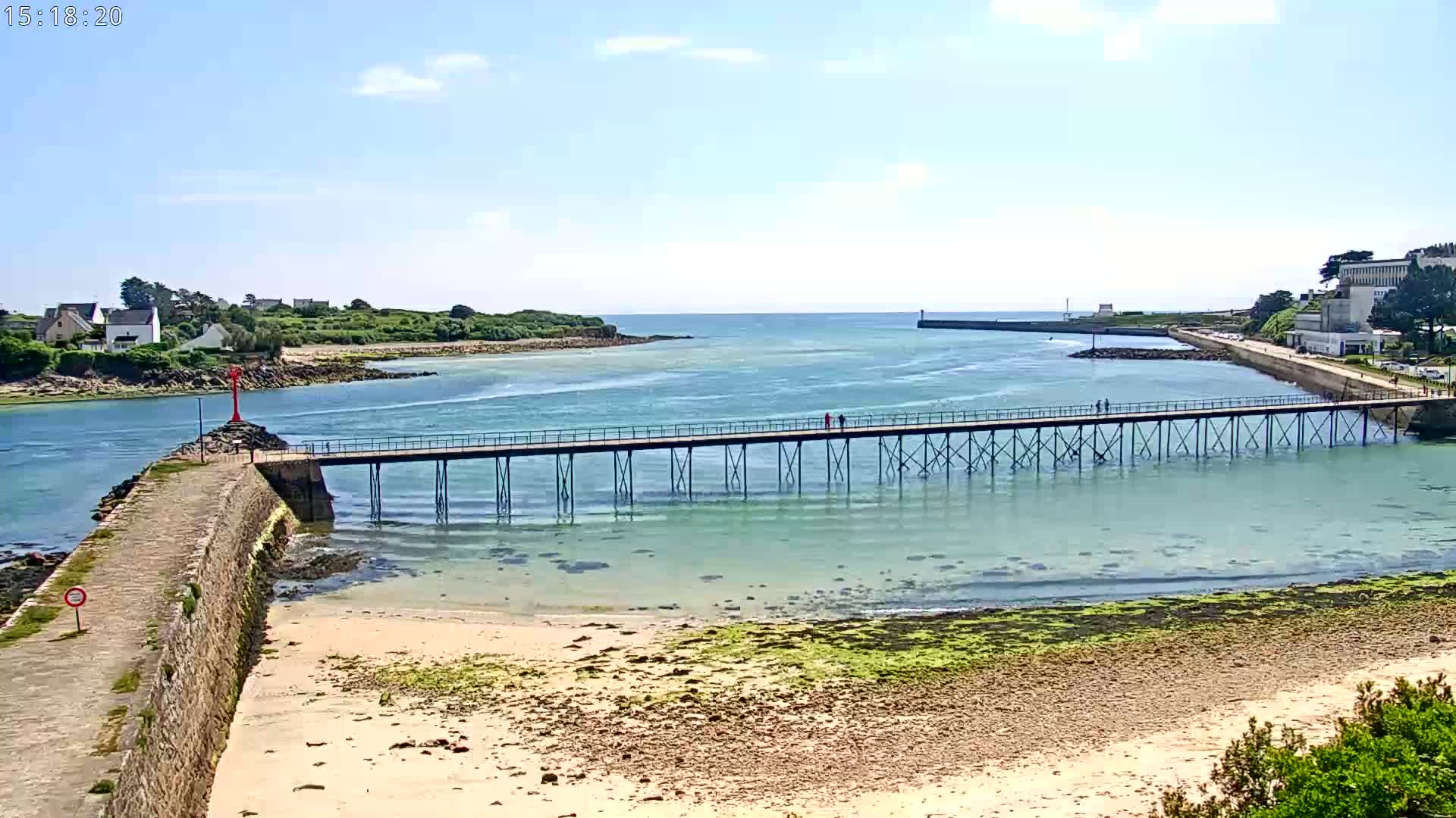 A long pier stretches across shallow, clear water to a breakwater on a sunny day, with a sandy beach and distant buildings visible.
