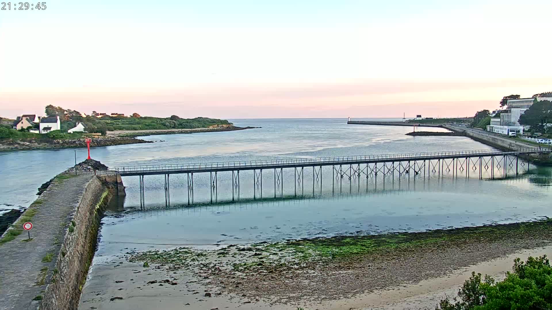 A long pier stretches across calm, shallow water at sunset, connecting two stretches of land with buildings and greenery visible in the distance.
