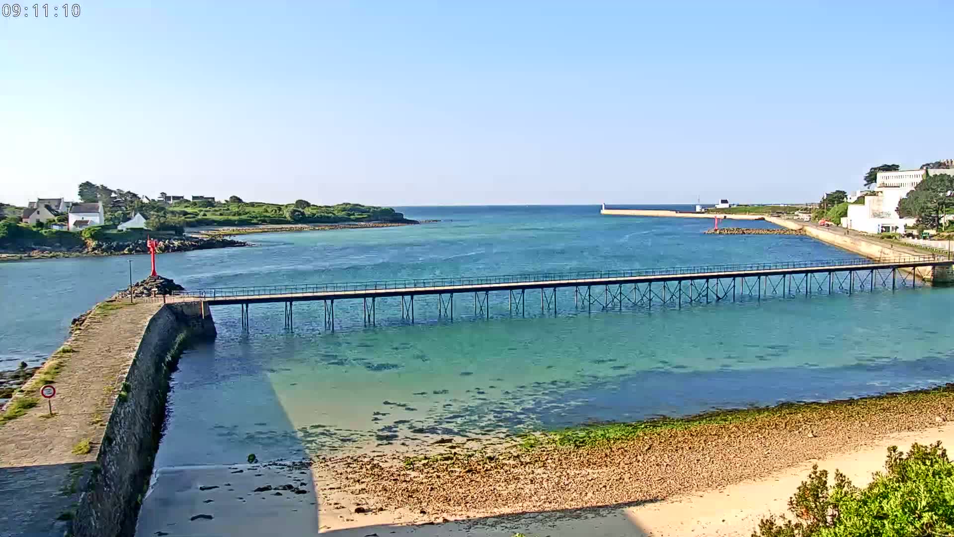 A long pier stretches across calm, clear turquoise water toward a small island and coastal buildings under a sunny sky.