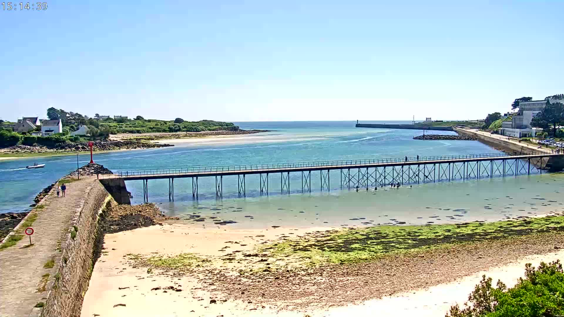 A long pier extends over shallow, clear water, connecting a stone wall to a landmass with buildings in the distance on a sunny day.