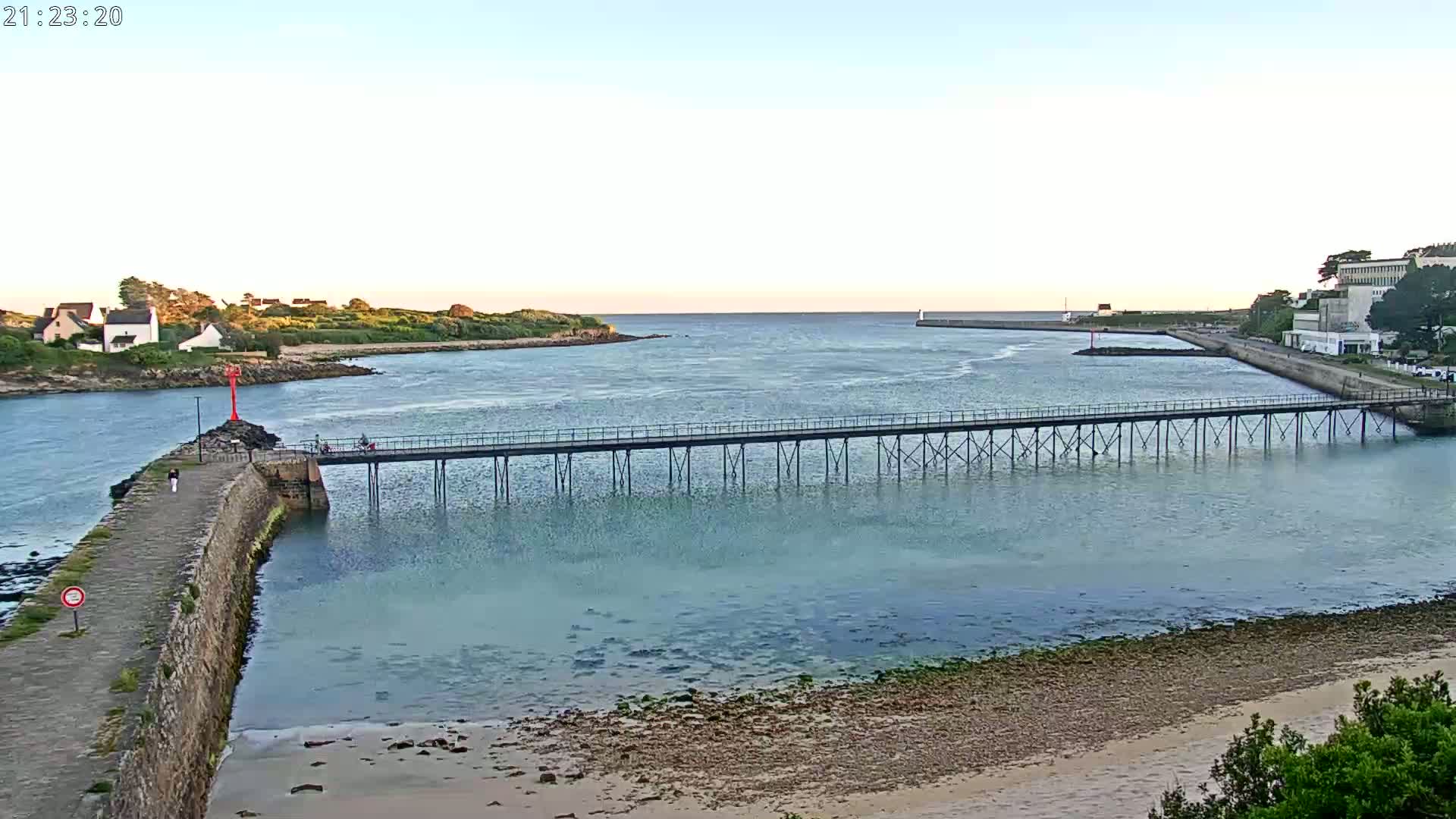 A long pier stretches across calm, shallow water separating a stone jetty from a shoreline with buildings and greenery under a pale evening sky.