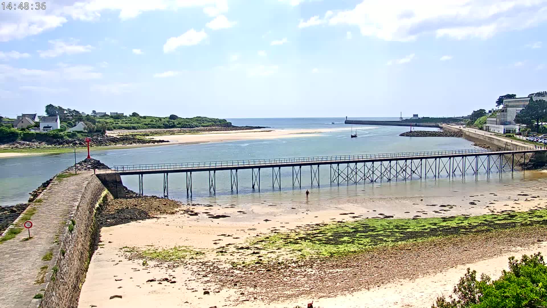 A long pier extends over shallow, sandy water towards a small island under a partly cloudy sky.