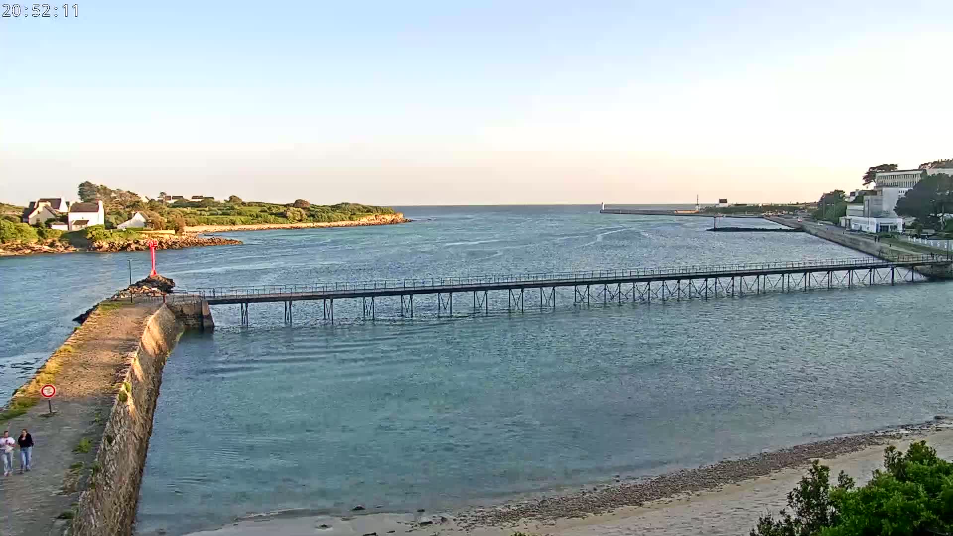A long pier stretches across calm, teal water, connecting a stone jetty to a shoreline with buildings and greenery under a clear, light-blue sky.