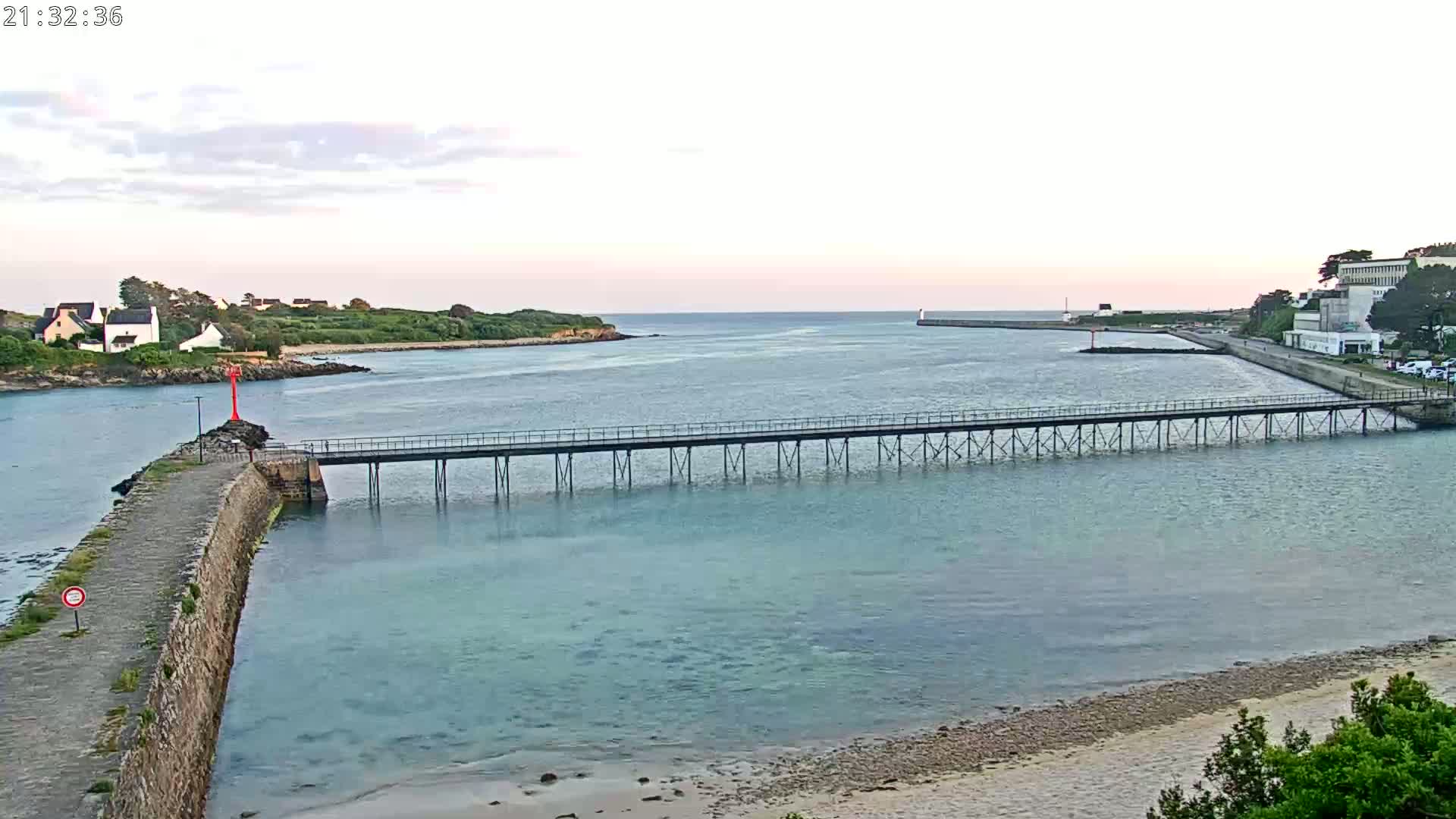 A long pier stretches across calm, light-blue water toward a small town and island under a pale, evening sky.