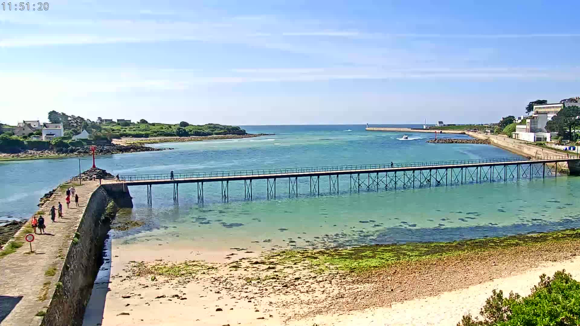 A long pier stretches across shallow, clear turquoise water to a breakwater, with people walking on a stone path and a sandy beach visible in the foreground under a sunny sky.