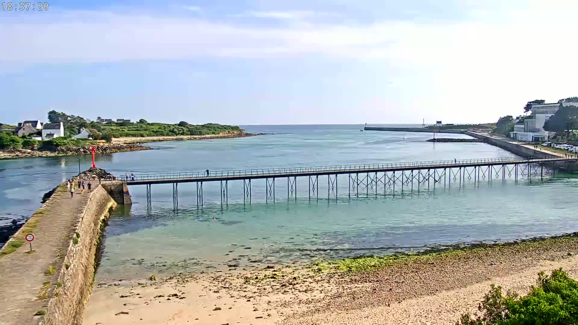 A long pier extends over calm, shallow, turquoise water toward a small island under a mostly sunny sky.